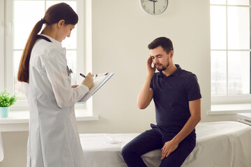 Young woman doctor nurse in uniform making notes during visit with man patient complaining of...