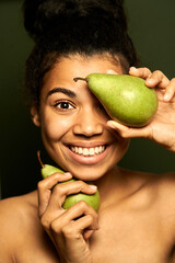 Choose fruits. Portrait of happy mixed race young woman smiling at camera, holding two juicy pears near her face, posing isolated over green background. Skincare, healthcare concept