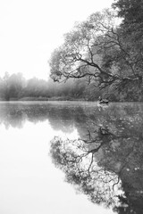 Pionowe czarno-białe ujęcie przedstawiające odbicie w tafli jeziora wędkarza na łódce i odbicie drzewa