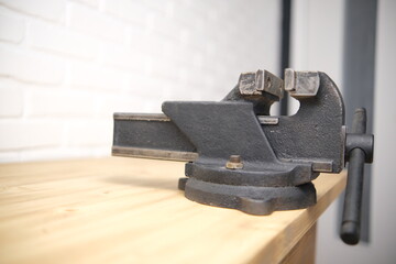 Close-up of a black vise with a screw grip and a cast-iron frame mounted on a wooden countertop, in a machine shop. The white brick background is blurred.