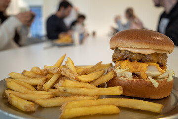Selective focused at cheese burger beef and fried potatoes, French fried on metal tray and white cafeteria table, and blur background of people eating and having lunch together in white canteen.  