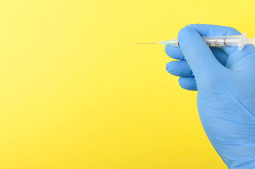 Hand wearing blue latex glove holding syringe on yellow background.