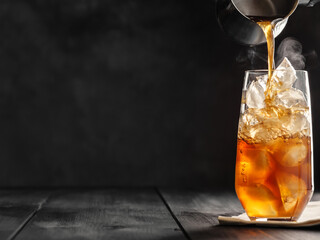 Cold coffee is poured from a Turk into a highball glass with ice on a gray wooden table. Iced coffee. Copy space, space for text.