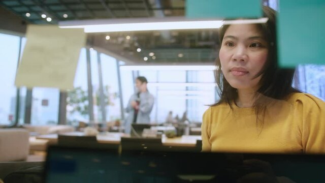 attractive freelance asian female creative person wear casual yellow cloth hand point and write down paper note stick reminder at coworking space new lifestyle with thoughful and freshness emotion