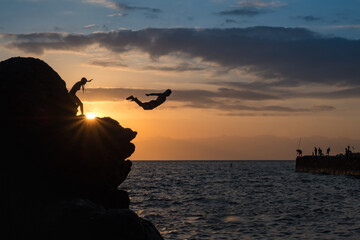 Young man jumping off cliff into ocean on sunset background. Business concept idea