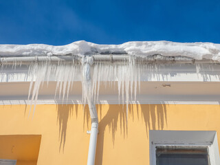 Large icicles under the roof of a yellow house on a sunny, clear day. Blue sky in the background