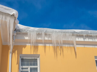 Large icicles under the roof of a yellow house on a sunny, clear day. Blue sky in the background
