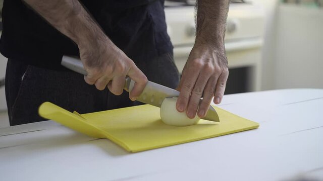 A man chopping onions with a sharp knife over a white table. Indoors, kitchen. 