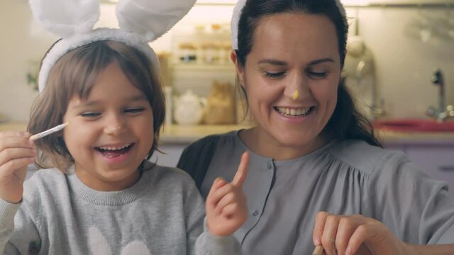 Happy Easter Holiday. Beautiful mother and daughter paint and decorate an egg for a traditional spring dinner at home. Girl with child playing, fooling, drawing easter eggs