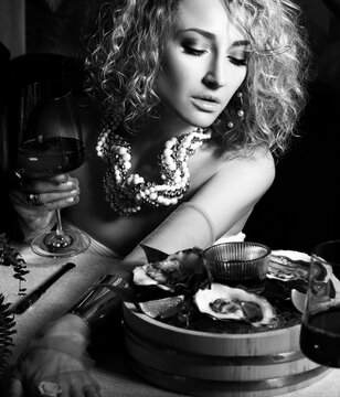 Black and white image of beautiful curly hair woman in seafood restaurant eating oysters and drink wine on dark background