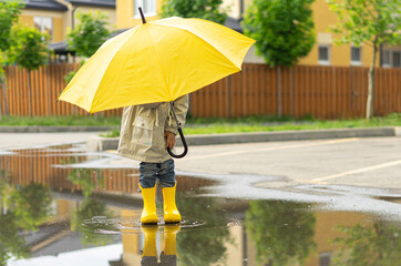 A child holds a yellow umbrella standing in a puddle outside. On sunny day happy kid walks near the...