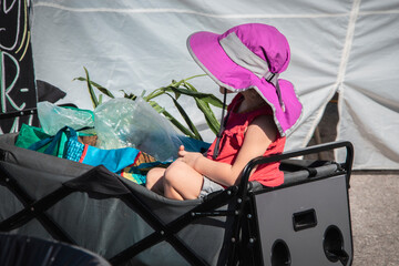 Little girl in fold-up wagon at outdoor market with plants and bags and bright pink sun hat hiding face