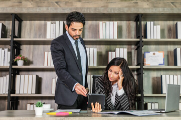 A businessman and businesswoman working together. The woman is sitting at her desk with her hand on her head while the man is standing next to her pointing to a report on tablet.
