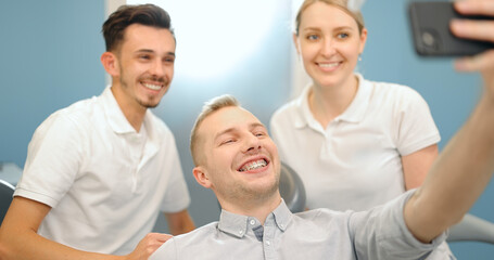 Obraz premium Young male patient in dental braces making selfie photo with a dentist and assistant during a regular orthodontic visit at the dental office. 4k video screenshot, please use in small size