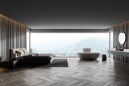 Master bedroom hotel room. Wooden walls, parquet floor. Comfortable king size bed standing on gray carpet. Bathtub near window with countryside view. Round mirror over double sink.