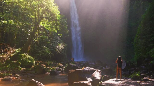 Silhouette beautiful woman enjoying amazing waterfall in the rays of the rising sun hidden in tropical rainforest jungle. Active lifestyle and Travel concept. 4K 