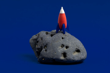 Cute miniature rocket on a moon stone on a dark blue background. Minimal space day card.