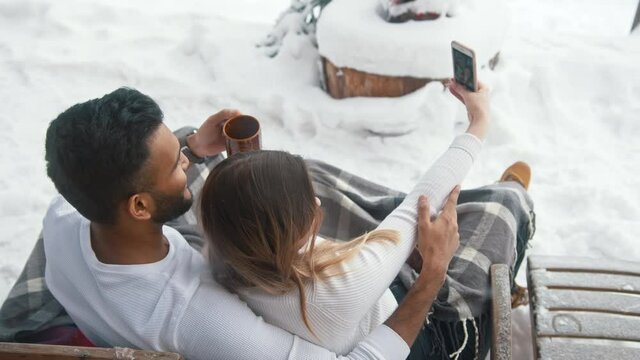 Multiracial couple sitting on the bench outdoors drinking hot chocolate and taking selfie on snowy valentines day. High quality 4k footage