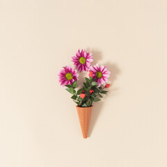 Ice cream cone with pink daisy flowers and green leaves on pastel beige background. Minimal summer flat lay composition.