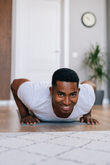 Fototapeta premium Low-angle shot of cheerful African-American man doing push-up on floor at cozy domestic room, looking at camera , close-up. Concept of sport workout training at home gym online in front of laptop .