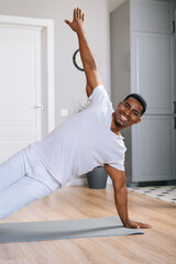 Cheerful sporty African-American man exercising in side plank position on floor during working out at bright domestic room. Concept of sport training at home gym.