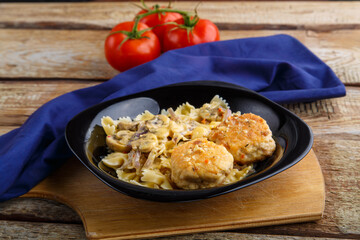 Pasta in a creamy sauce with mushrooms and chicken meatballs in a plate on a board on a blue napkin next to a tomato.