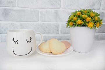 a cup with a funny face, a plate with cookies and hearts and an artificial flower in a flowerpot on the table in the kitchen.
