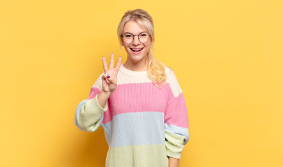 blonde woman smiling and looking friendly, showing number three or third with hand forward,...