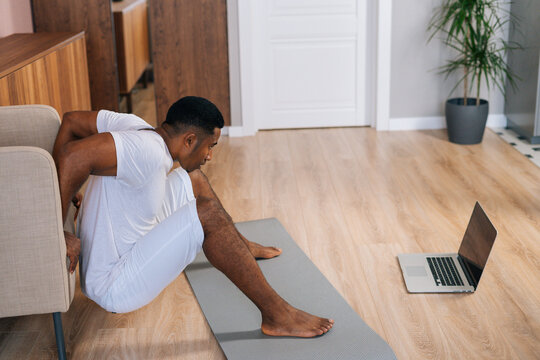 Young African-American man doing reverse push-up online in front of laptop for triceps muscles using armchair at bright domestic room. Concept of sport workout training at home gym.