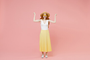 Full length body young cute happy excited redhead woman in straw hat glasses summer clothes maxi skirt do winner gesture clenching fists look camera isolated on pastel pink background studio portrait