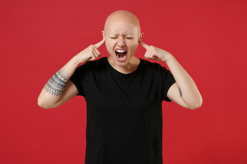 Fototapeta premium Young bald caucasian tired stressed tattooed woman 20s without hair wearing black basic casual t-shirt covering ear with fingers hands shout scream isolated on red color background studio portrait.