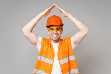 Young employee man in orange vest protective hardhat hold folded hands above head like roof stay home isolated on grey background studio Instruments for renovation apartment room. Repair home concept.