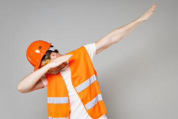 Young employee smiling man in orange vest protective helmet hardhat do dab hip hop dance hands move gesture isolated on grey background studio Instruments for renovation apartment Repair home concept