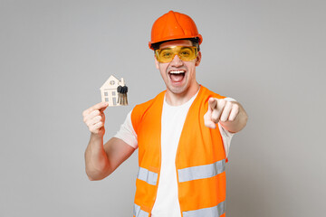 Young man in orange vest protective hardhat hold house bunch of keys point index finger camera on you isolated on grey wall background studio Instruments for renovation apartment Repair home concept.
