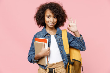Happy little african american kid school girl 12-13 years old in casual denim clothes backpack hold books show ok okay gesture isolated on pastel pink background studio Childhood education concept.