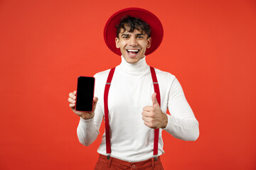 Young spanish latinos smiling satisfied happy man 20s wearing hat, white shirthold mobile cell phone with blank screen workspace area show thumb up gesture isolated on red background studio portrait.