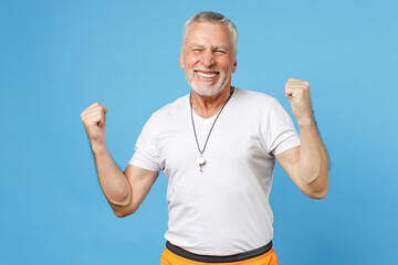 Fototapeta premium Elderly gray-haired sportsman trainer instructor strong man 50s in sportswear white t-shirt whistle do winner gesture clench fist celebrating isolated on blue background studio. Fitness sport concept