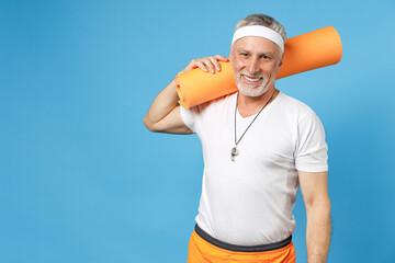 Elderly gray-haired sportsman rainer instructor smiling coach man 50s wearing sportswear white t-shirt holding yoga mat on shoulder isolated on blue background studio portrait. Fitness sport concept