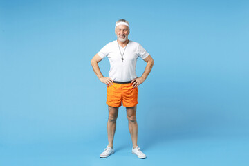 Full length elderly gray-haired sportsman fitness instructor smiling man 60s in sportswear white t-shirt whistle stand akimbo arm on waist isolated on blue background studio portrait. Sport concept.