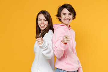 Cheerful smiling funny two young women friends 20s wearing casual white pink hoodies standing back to back pointing index fingers on camera isolated on bright yellow color background studio portrait.
