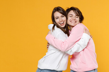 Smiling cheerful two young women friends 20s wearing casual basic white pink hoodies standing hugging having fun looking camera keeping eyes closed isolated on yellow color background studio portrait.