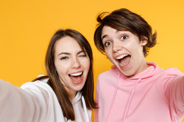 Close up of excited blinking cheerful two young women friends 20s wearing casual basic white pink hoodies doing selfie shot on mobile phone isolated on bright yellow color background studio portrait.