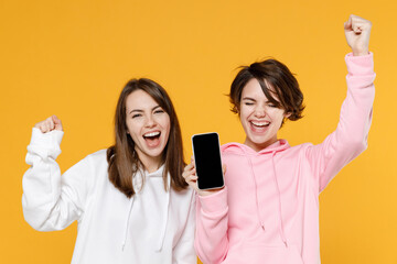 Happy two young women friends 20s wearing casual white pink hoodies hold mobile cell phone with...