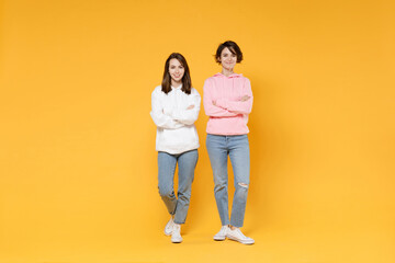 Full length of smiling attractive two young women friends 20s wearing basic white pink hoodies standing holding hands crossed looking camera isolated on bright yellow color background studio portrait.