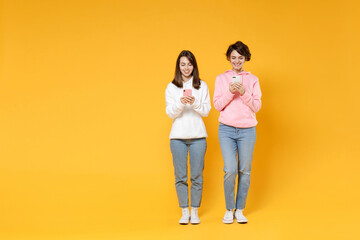 Full length of smiling attractive two young women friends 20s wearing casual white pink hoodies using mobile cell phone typing sms message isolated on bright yellow color background studio portrait.