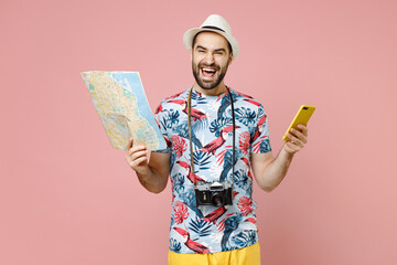Laughing young traveler tourist man in summer clothes hat using mobile cell phone hold city map isolated on pink background. Passenger traveling abroad on weekends getaway. Air flight journey concept.