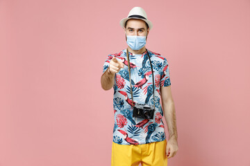 Funny traveler tourist man in face mask to safe from coronavirus virus covid-19 point index finger camera on youn isolated on pink background. Passenger travel on weekend. Air flight journey concept.