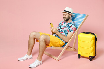 Full length smiling young traveler tourist man in hat sit on deck chair using mobile cell phone...