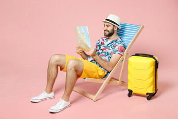Full length of smiling young traveler tourist man in summer clothes hat sit on deck chair hold city...