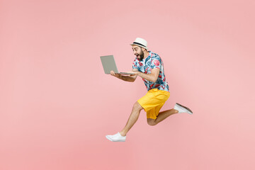 Full length side view of excited traveler tourist man in summer clothes hat jumping working on laptop computer isolated on pink background. Passenger traveling on weekend. Air flight journey concept.
